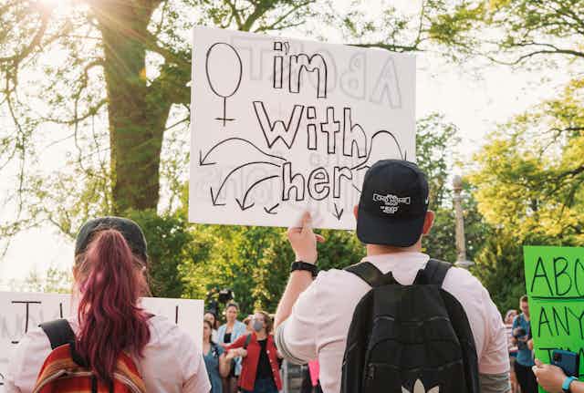 A man holds a sign at a protest reading "I'm with her".