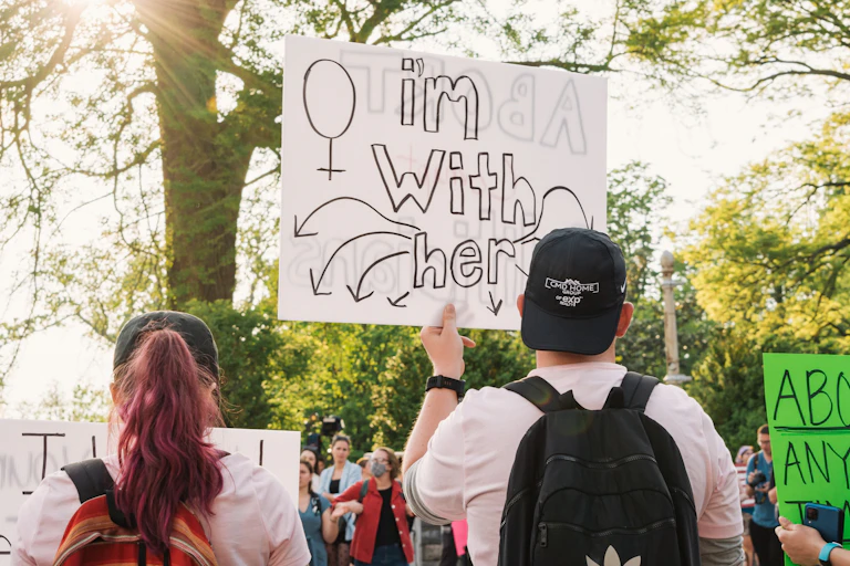 A man holds a sign at a protest reading "I'm with her".