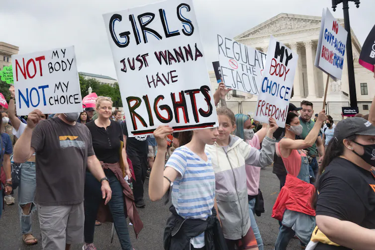 A male protestor holds a sign reading 'not my body, not my choice' and another protestor holds a sign reading 'girls just wanna have rights'.
