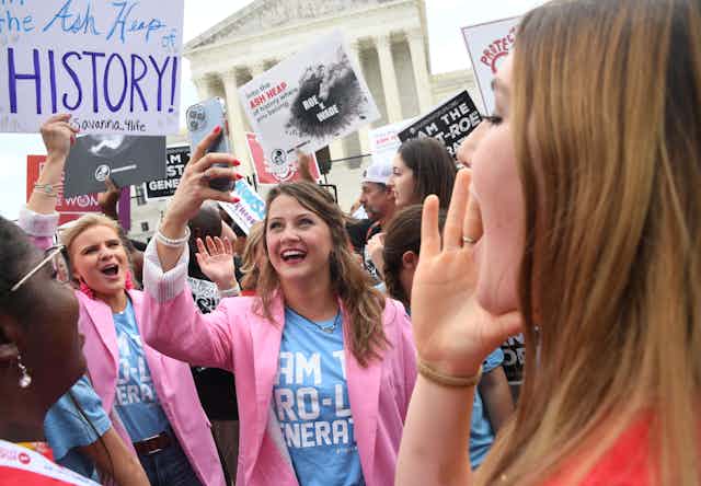 Young women cheer and smile and hold signs that say things like 'the ash heap of history' outside the Supreme Court