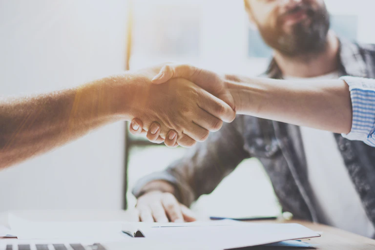 Handshake between two people, two more in the background, laptop in the foreground