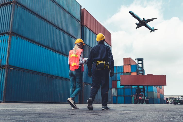 Two people walking past storage containers wear hard hats and high-visibility clothing, plane flying overhead.