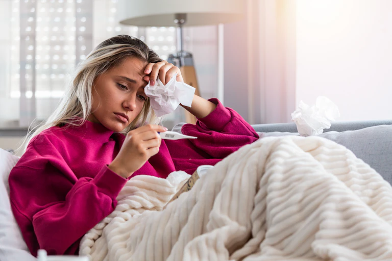 A young woman lying on a couch looks at a thermometer.