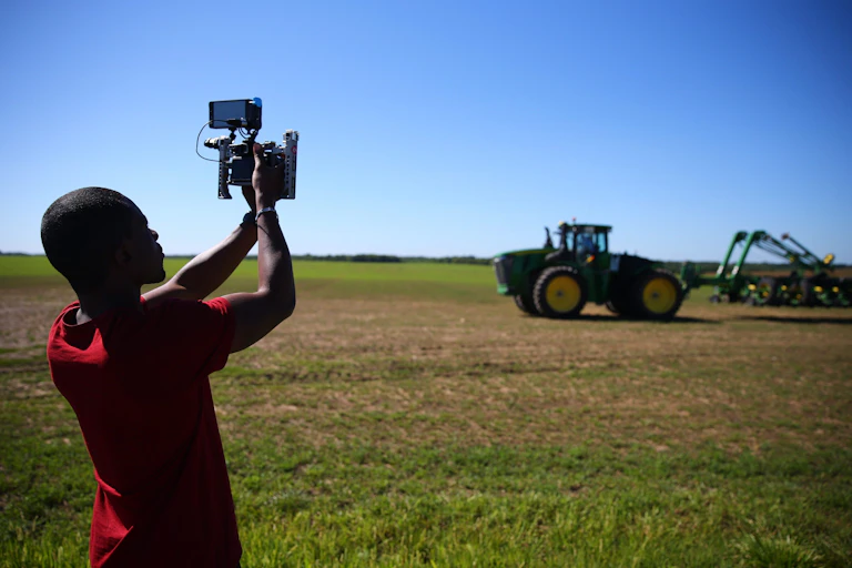 A man holds up a camera phone attached to equipment pointed at a tractor on farmland