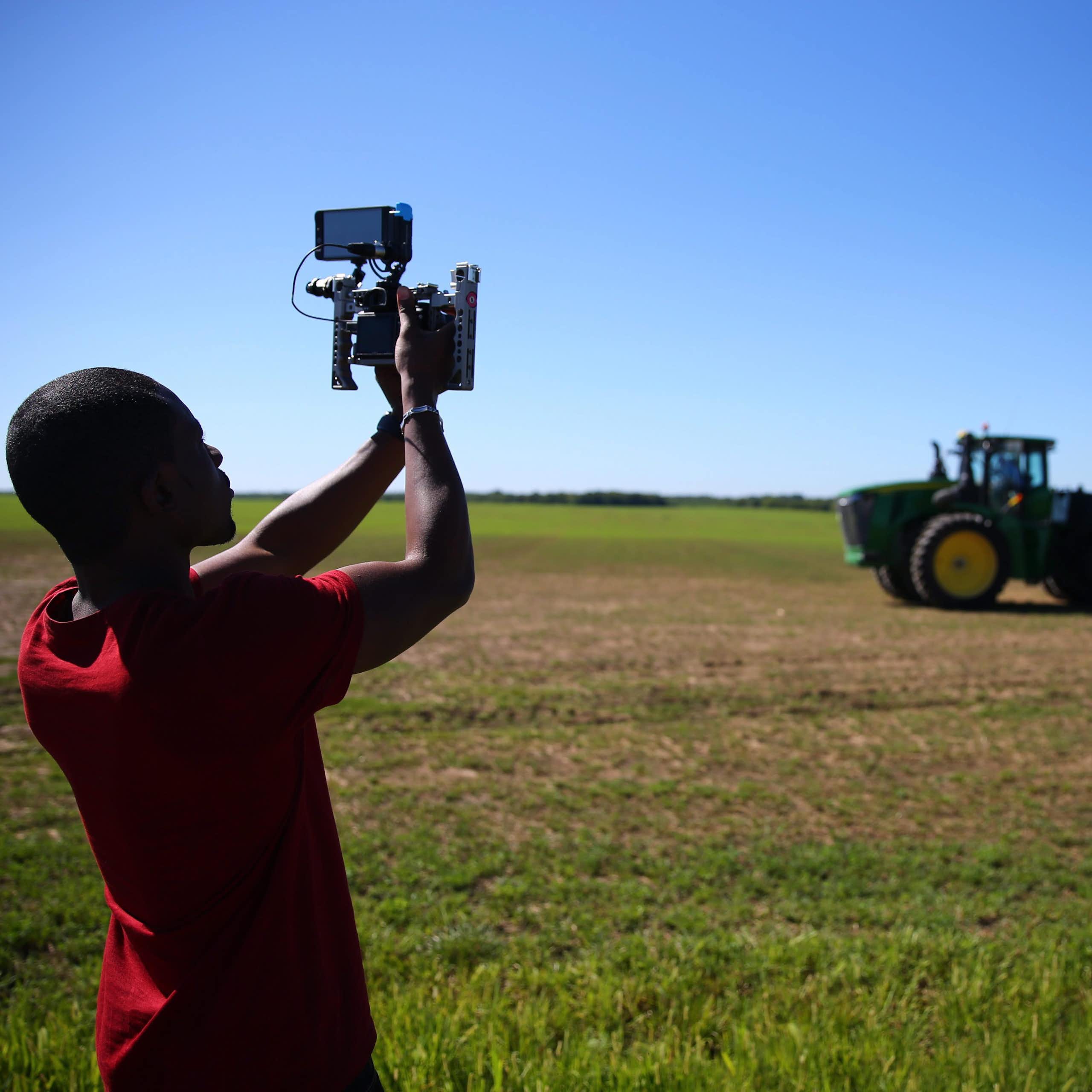 A man holds up a camera phone attached to equipment pointed at a tractor on farmland