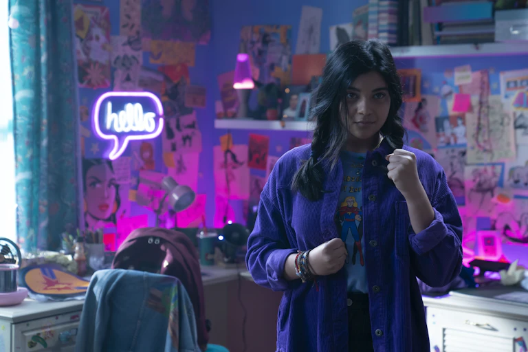 A young woman in her room surrounded by purple-hued walls and art and comic pictures, seen clenching one fist near her body and holding one up, looking forward.