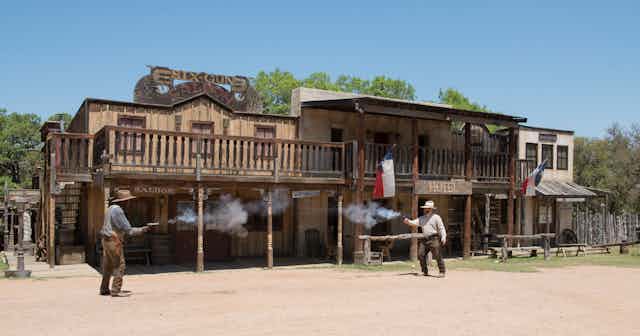 Two men fire pistols at each other in front of a row of buildings