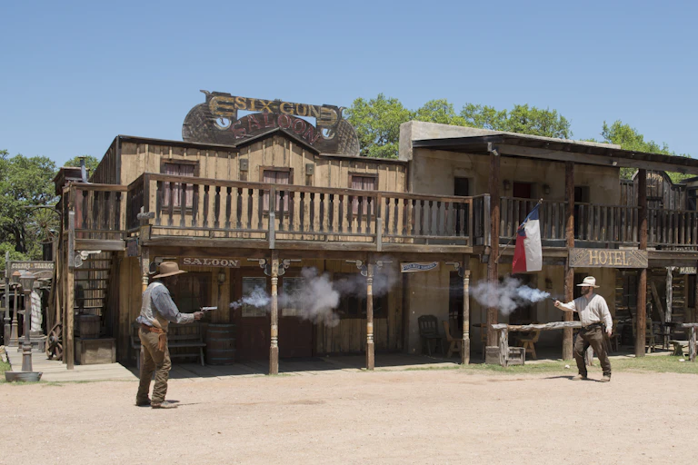 Two men fire pistols at each other in front of a row of buildings