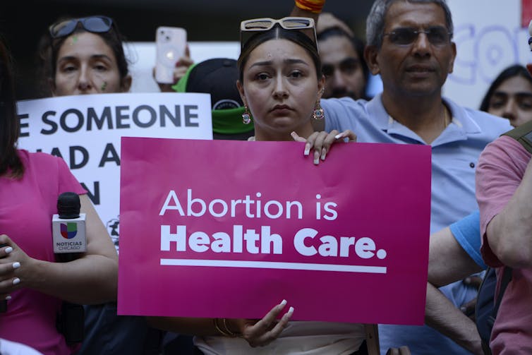A woman in a crowd of protesters holding a bright pink sign with white lettering reading 'Abortion is health care'