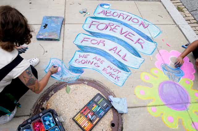 A person seen from behind with art supplies colouring a banner on the sidewalk reading 'Abortion forever for all we are never going back'