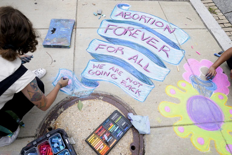 A person seen from behind with art supplies colouring a banner on the sidewalk reading 'Abortion forever for all we are never going back'