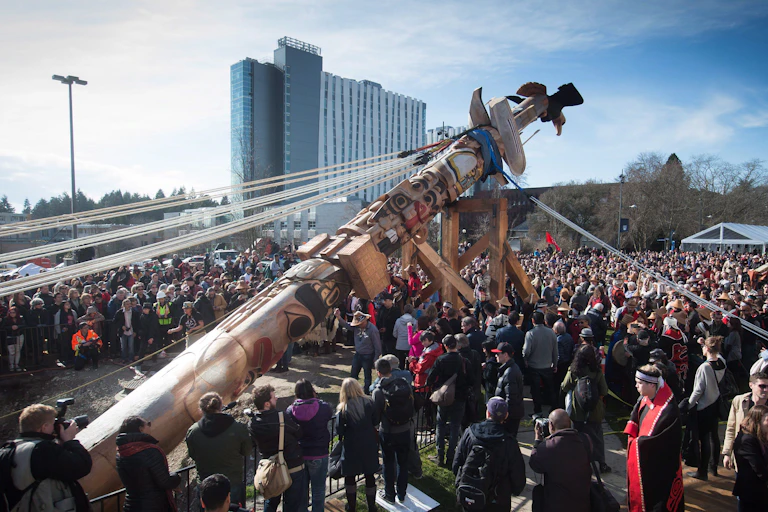 People are seen in a crowd in front of a tall modern apartment-style building surrounding a totem pole with a large bird on the top tilted on its side being held by cords; the pole looks like it is going to be raised.