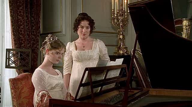 a young woman sits at a piano, with another young woman standing beside her