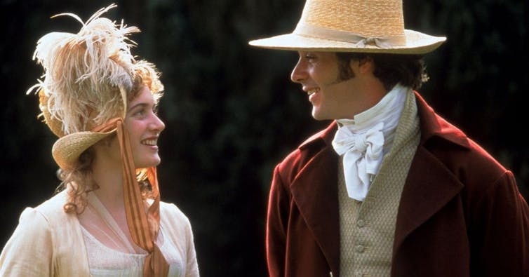 A woman in a feathered hat smiles at a man in a straw hat.