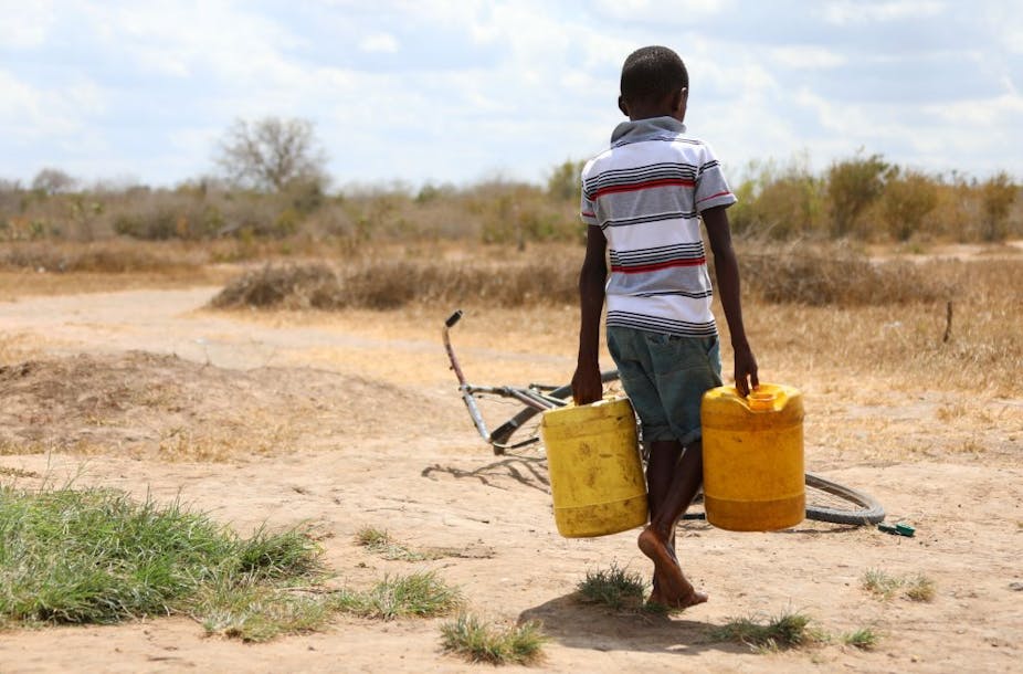 A boy carrying two jerricans of water in a dry field.