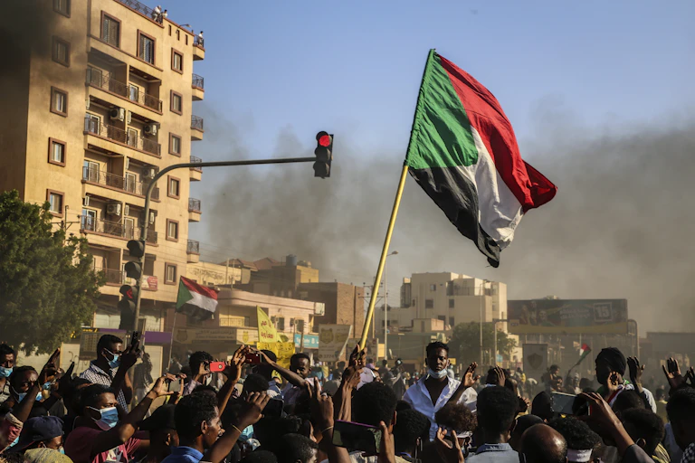 Hundreds of youthful protesters carry the Sudanese flag on sticks.