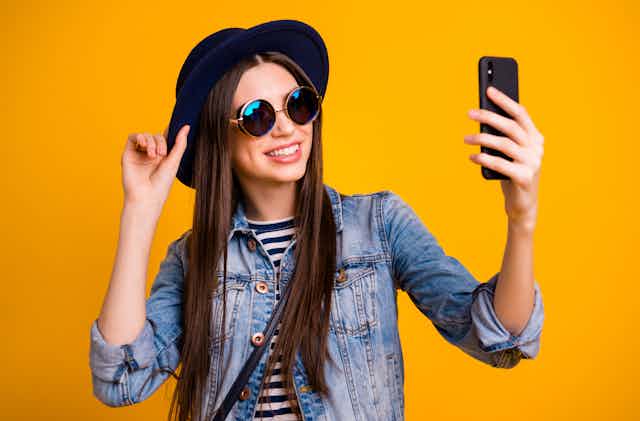 A woman in a stylish hat and sunglasses takes a selfie against a yellow background