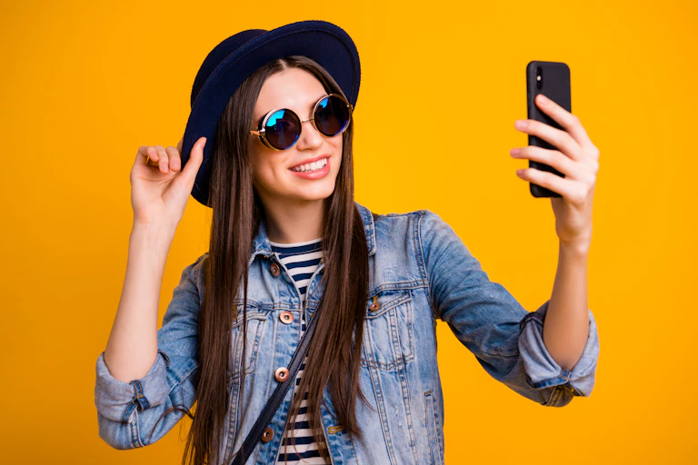 A woman in a stylish hat and sunglasses takes a selfie against a yellow background