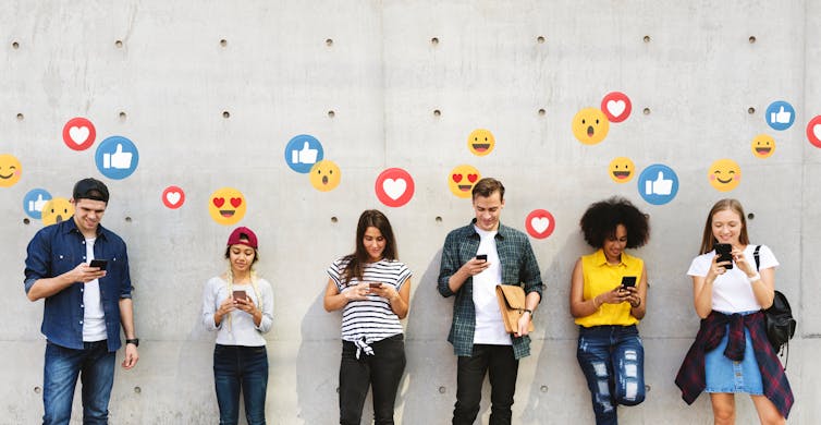 Six young people leaning against a concrete wall, all watching their mobile phones. Emoji icons are emanating from the screens.