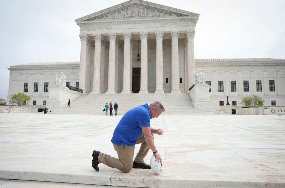 A man in a blue shirt and khaki-colored pants kneels, clutching a football, in front of the US Supreme Court.