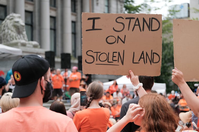 A woman holds a sign that reads 'I stand on stolen land'.