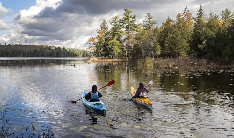Two women kayak in a blue and yellow kayak on a lake with trees in the background