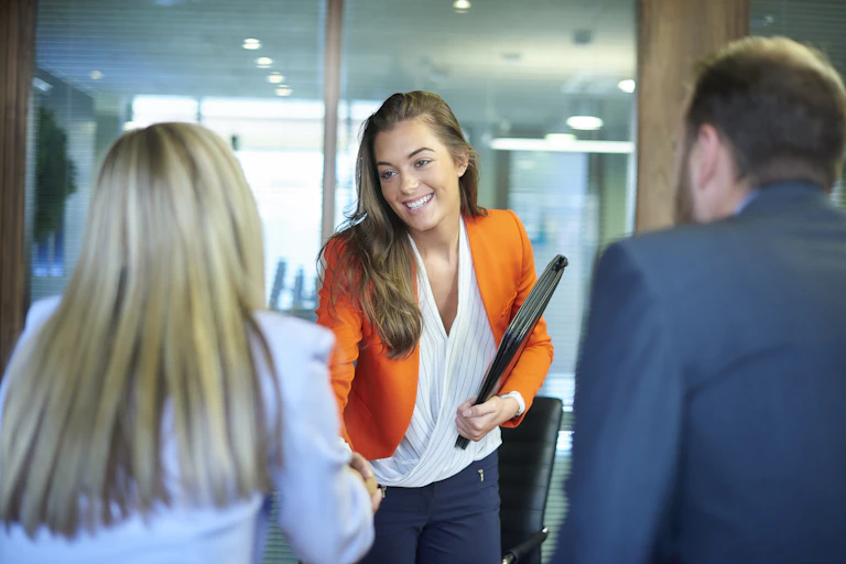 A young woman in an orange blazer shakes another woman's hand in an office while a man in a blue suit jacket stands nearby.