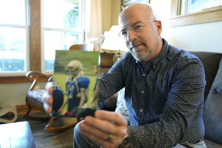 A man in a blue shirt sits as he holds up a color photograph of a young man in a football uniform.