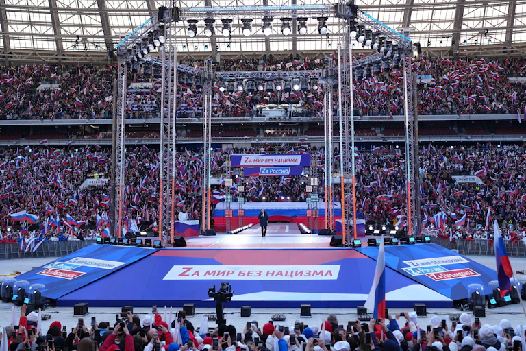 A stadium shows many people around a stage Putin stands on.