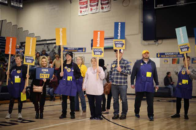 Seven people in a line holding signs for Pete Buttigieg, a Democratic presidential candidate, in a large gym.