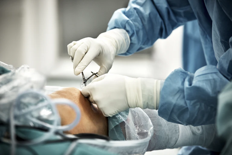 A surgeon's gloved hands are holding medical scissors while sewing up a patient