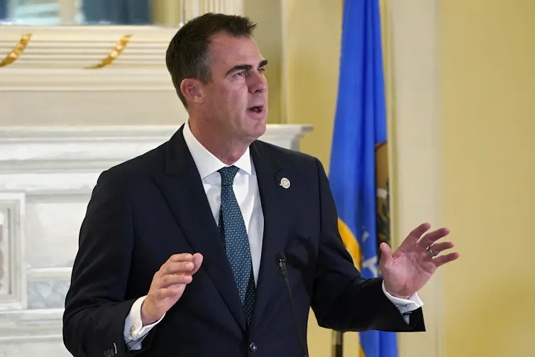 A man in a blue suit stands in front of a mantle with his hands up