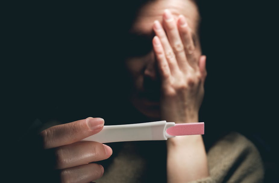 A woman viewing a positive pregnancy test covers one eye with her hand in fear.