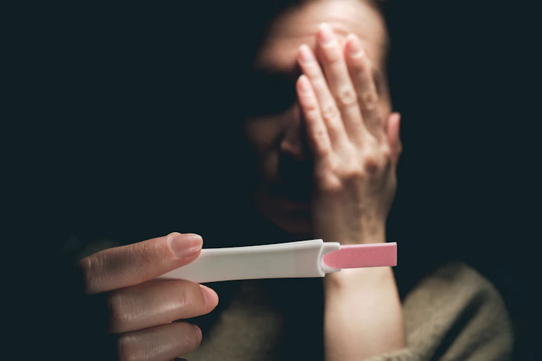 A woman viewing a positive pregnancy test covers one eye with her hand in fear.