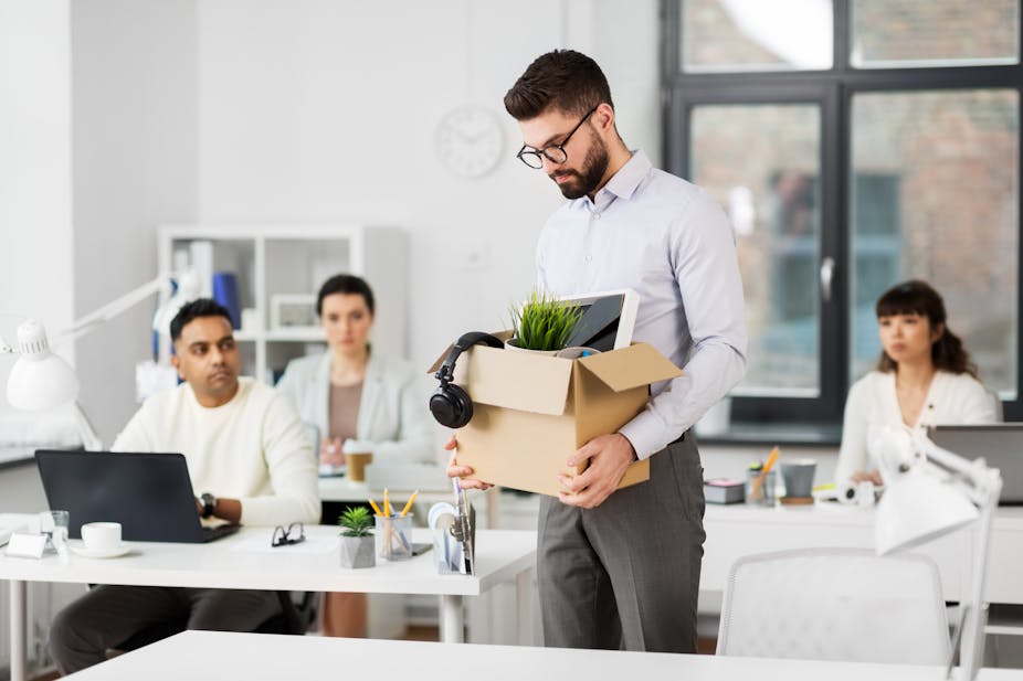 Man with box of belongings in office, co-workers in the background.