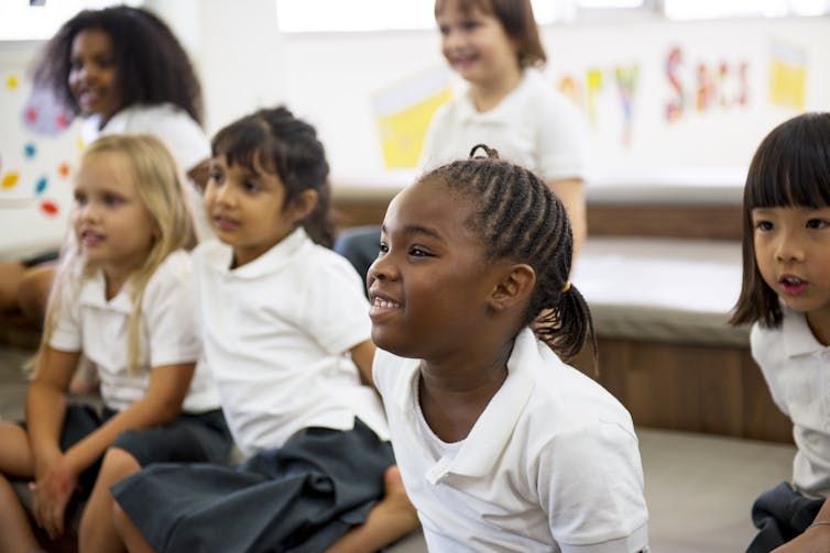 Primary school children in uniform sit on the ground looking in the same direction.