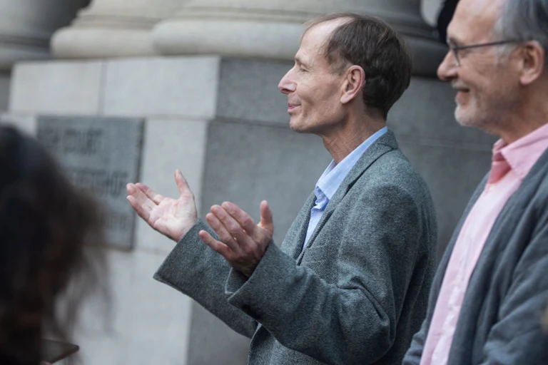 Men in grey suits in profile outside a stone building. One, balding, holds up his hands, palms upward.