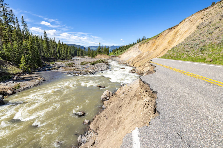 A road ends in a jagged cliff where the swollen river had raced through and scoured far up the hillside where the rest of the road once existed. Evergreens and mountains are in the background on a sunny day.