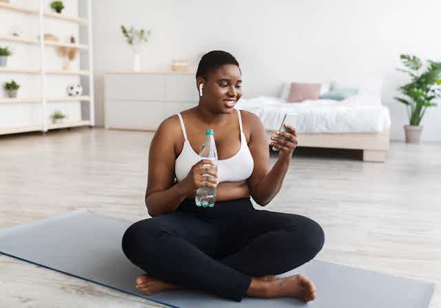 Woman in fitness clothes sitting cross-legged on a yoga mat holding a bottle of water and looking at a smartphone