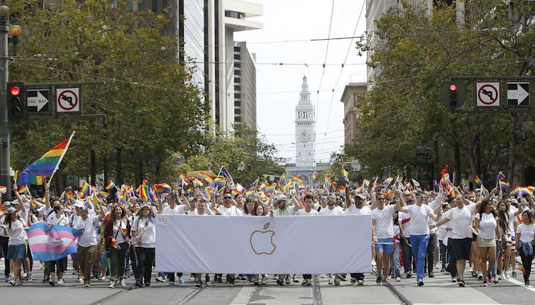 a crowd of people holding a large white banner with an apple outlined on it march on a street carrying rainbow flags