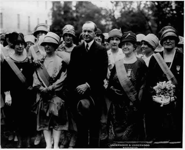 Calvin Coolidge stands in a row of women wearing sashes in this black and white photo