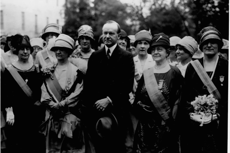 Calvin Coolidge stands in a row of women wearing sashes in this black and white photo