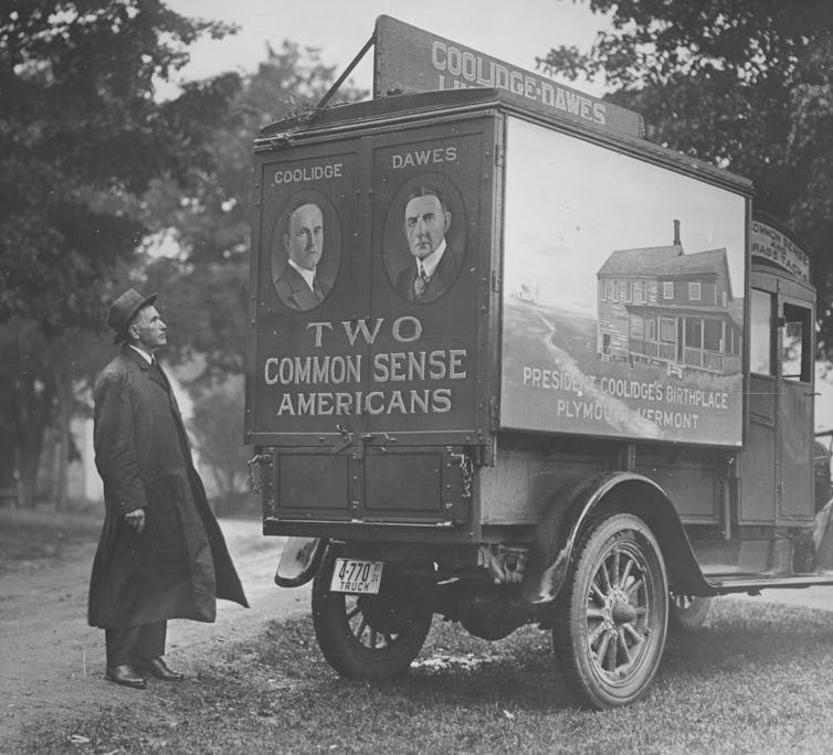 A black and white photo depicts a man in a topcoat and hat gazing at a truck bearing images of two men and the words 'Two common sense Americans.'