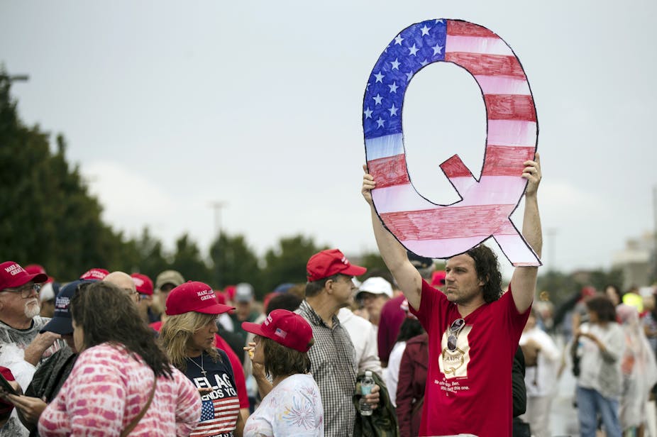 A man in a red shirt holds up a large letter Q in the colors of American flag in a crowd of people wearing red hats.