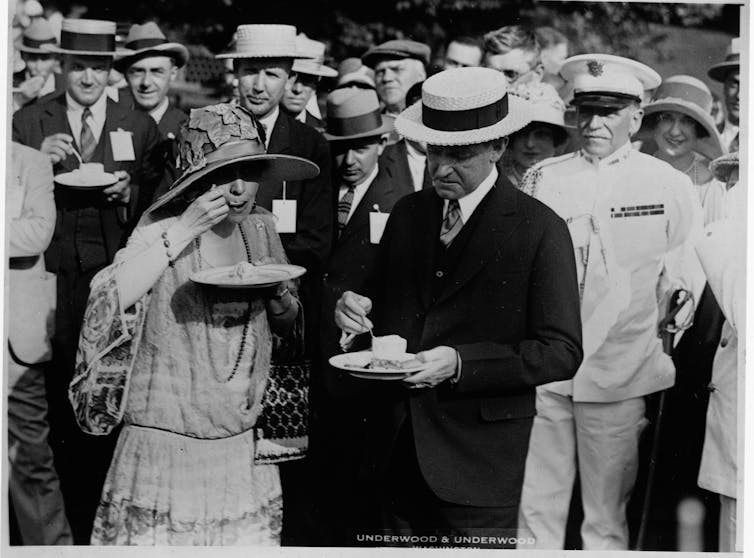 Calvin Coolidge eats ice cream off a plate next to his wife, in front of a group of men dressed formally in suits and a Navy uniform in this black and white photo