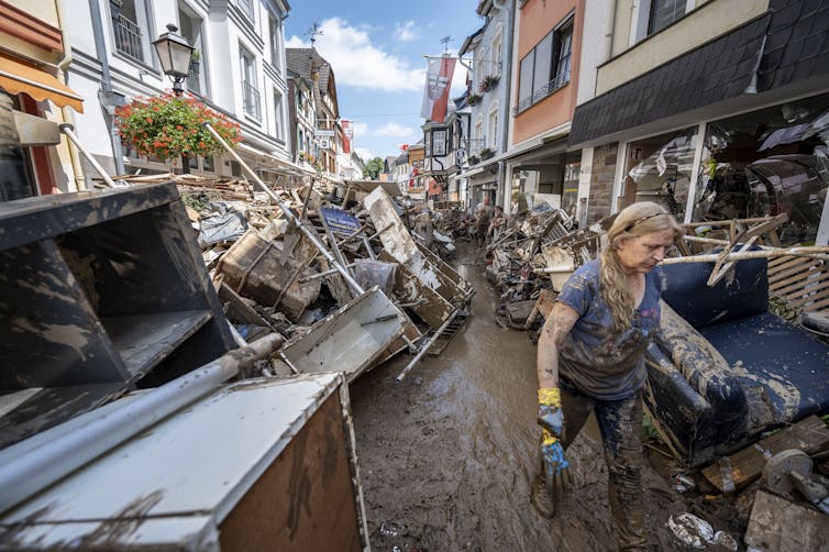 A woman with work gloves and clothing covered in mud walks through a muddy residential street filled with mud-covered furniture and other damaged belongings people are throwing out after a flood.