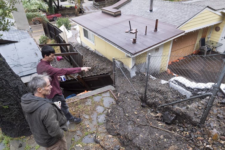 Two men point stand on a deck overlooking a neighboring house where mud has flowed through the yard and is mounded half way up the side of the home.