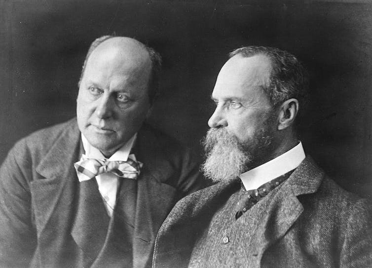 A black and white photograph shows two men posed next to each other in suits.