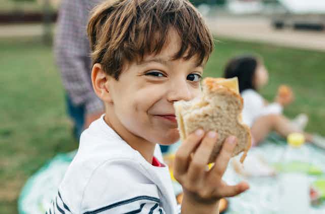 A grinning boy holds up half a sandwich with a bite out of it. A picnic blanket and family are in the background.