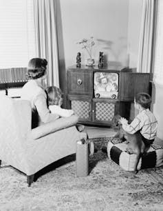 A black-and-white photograph of a mother and her two children watching television in the living room.